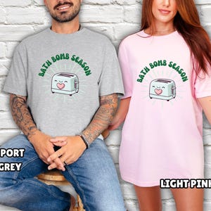 Puede incluir: Una camiseta gris claro y una camiseta rosa claro, cada una con el texto "BATH BOMB SEASON" encima de una tostadora de dibujos animados con un coraz&oacute;n. La camiseta gris est&aacute; etiquetada como "SPORT GREY" y la camiseta rosa como "LIGHT PINK".