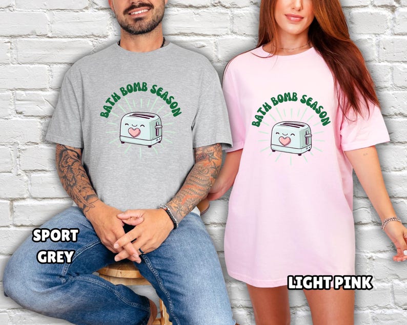 Puede incluir: Una camiseta gris claro y una camiseta rosa claro, cada una con el texto "BATH BOMB SEASON" encima de una tostadora de dibujos animados con un coraz&oacute;n. La camiseta gris est&aacute; etiquetada como "SPORT GREY" y la camiseta rosa como "LIGHT PINK".