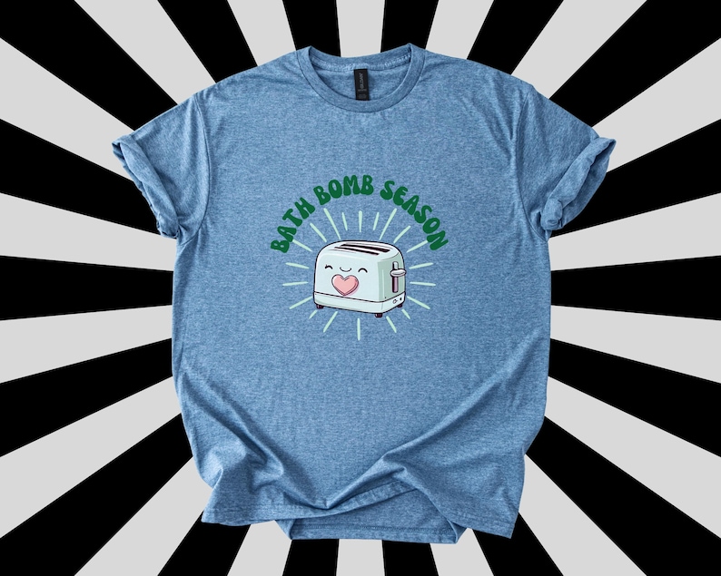 Puede incluir: Camiseta azul claro con mangas enrolladas. La camiseta presenta una tostadora de dibujos animados con un coraz&oacute;n y el texto "BATH BOMB SEASON" en verde, rodeado de l&iacute;neas radiantes. El fondo es un patr&oacute;n de rayas blancas y negras.
