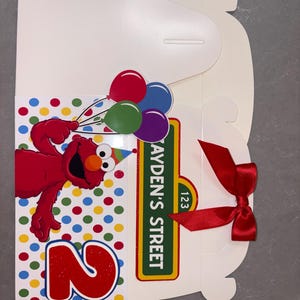 May include: A white birthday gift box featuring Elmo holding balloons. The box has a colorful polka dot pattern and a red number 2. The box is tied with a red ribbon and has a street sign that reads "AYDEN'S STREET 123".
