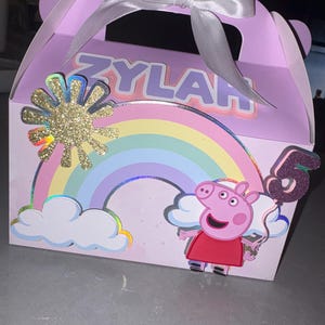 May include: A lavender party favor box with a white ribbon bow. The box features a rainbow, a glittery sun, and a cartoon pig holding a number 5 balloon. The name "ZYLAH" is displayed on the box.