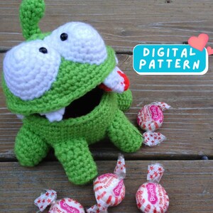 May include: Green crochet monster with a big mouth and white eyes. The monster is holding a candy in its mouth and there are several candies on the wooden surface. The text "DIGITAL PATTERN" is on a blue background with pink hearts.