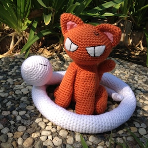 May include: A crocheted orange cat with a white crocheted snake wrapped around it. The cat has a toothy grin and is sitting on a white surface.