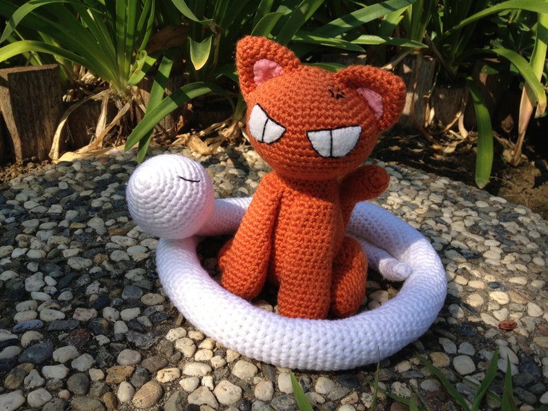May include: A crocheted orange cat with a white crocheted snake wrapped around it. The cat has a toothy grin and is sitting on a white surface.