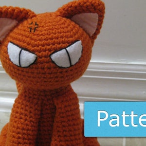 May include: Orange crocheted cat plush toy with white eyes and black pupils. The cat has a grumpy expression and is sitting on a white surface.