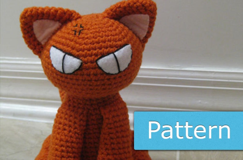 May include: Orange crocheted cat plush toy with white eyes and black pupils. The cat has a grumpy expression and is sitting on a white surface.