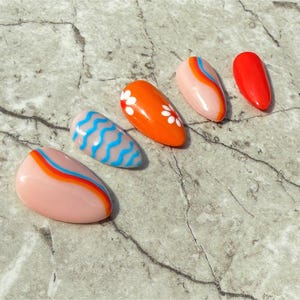May include: Five almond-shaped artificial nails with colorful designs. One nail is solid red, while others feature patterns like wavy blue lines, orange with white flowers, and pink with orange and blue stripes. The nails are arranged on a textured gray surface.