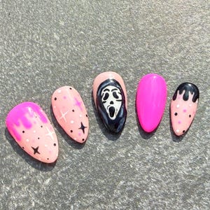 May include: Set of five artificial nails in various designs. The nails feature a pink and black color scheme with a Scream mask design, polka dots, and star accents. One nail is solid bright pink.