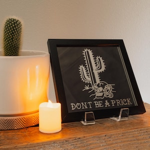 May include: A black framed print with a cactus and skull design, with the text "DON'T BE A PRICK". A cactus in a white pot and two small, lit candles are also on the wooden surface.