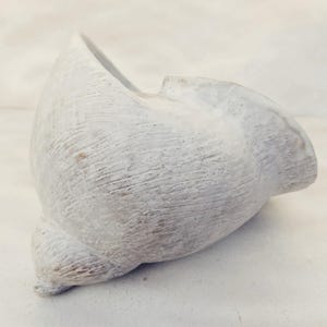 Op de afbeelding: Een witte keramische kom met een getextureerd, verweerd buitenoppervlak met fijne, geëtste lijnen en een gladde, glanzende binnenafwerking. Het vat heeft een onregelmatige, organische vorm met een brede, uitlopende rand en een taps toelopende basis.