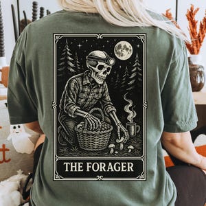 May include: Olive green t-shirt featuring a detailed graphic of a skeleton foraging in a forest under a full moon. The graphic includes the text "THE FORAGER" in a decorative frame. The shirt is a relaxed fit.