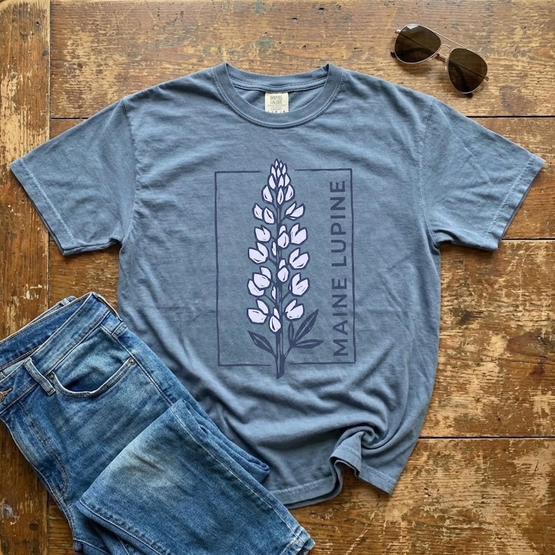 May include: A blue t-shirt with a Maine Lupine design. The graphic features a purple lupine flower within a rectangular frame, with the words "MAINE LUPINE" printed vertically. The shirt is paired with blue jeans and sunglasses.