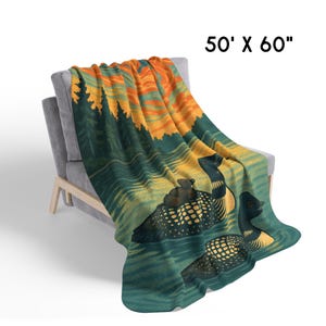 Może przedstawiać: Koc o wymiarach 127 cm x 152 cm narzucony na szary fotel. Koc przedstawia scenę przyrodniczą z dwoma nurami na jeziorze, zachodem słońca i drzewami. Nury są czarne i ż&oacute;łte, a woda turkusowa.