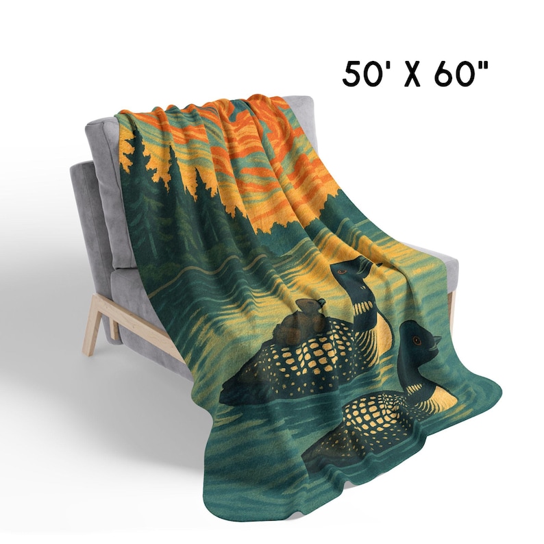 Może przedstawiać: Koc o wymiarach 127 cm x 152 cm narzucony na szary fotel. Koc przedstawia scenę przyrodniczą z dwoma nurami na jeziorze, zachodem słońca i drzewami. Nury są czarne i ż&oacute;łte, a woda turkusowa.