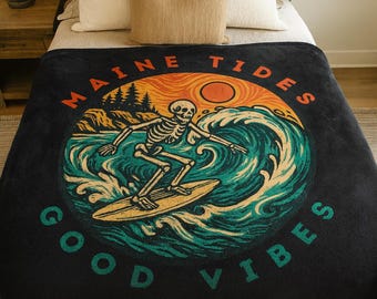 May include: Black blanket with a circular graphic featuring a skeleton surfing a wave. The design includes the text "MAINE TIDES" and "GOOD VIBES" in orange and teal. The background shows a sunset and trees.