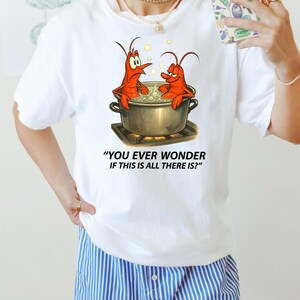 May include: White t-shirt with a cartoon image of two red lobsters in a cooking pot. The text reads "YOU EVER WONDER IF THIS IS ALL THERE IS?". A humorous design, perfect for casual wear. The shirt is white.