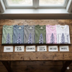 May include: Six folded t-shirts in various colors, including bay, blue jean, granite, light green, orchid, and white. Each shirt features a floral design with the text "JE LUPINE" in a rectangular frame. The shirts are displayed on a wooden surface.