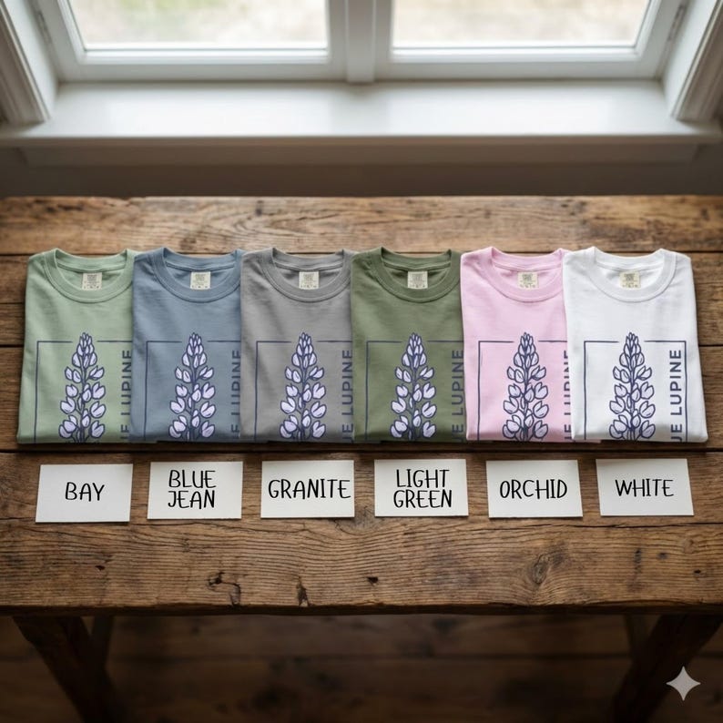 May include: Six folded t-shirts in various colors, including bay, blue jean, granite, light green, orchid, and white. Each shirt features a floral design with the text "JE LUPINE" in a rectangular frame. The shirts are displayed on a wooden surface.