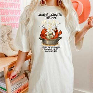 May include: A cream-coloured t-shirt with the words "MAINE LOBSTER THERAPY" and a cartoon of two lobsters in a pot. Below the image is the text "LOOK! WE'RE FINALLY WARMING UP TO EACH OTHER!". The shirt is a relaxed fit.