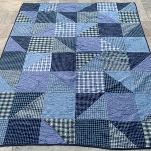 May include: A patchwork quilt in shades of blue, featuring squares and triangles of various patterns, including solid, gingham, and textured fabrics. The quilt is laid out on a tiled surface, showcasing its geometric design and color palette.