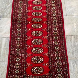 May include: A rectangular red rug with an intricate geometric pattern. The rug features a central design of repeating medallions in shades of red, brown, and cream, bordered by detailed patterns. The rug's edges are finished with a fringe.