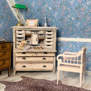 Puede incluir: Un escritorio secreter en miniatura de madera con acabado blanco desgastado, que presenta múltiples cajones pequeños, una superficie de escritura abatible y tres cajones grandes. Un sillón rosa pequeño y una alfombra de leopardo están delante.