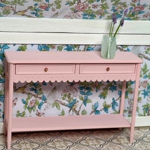 May include: A light pink, two-drawer console table with a scalloped edge and a lower shelf. The table has brass knobs and is topped with a small glass jar holding purple flowers. The background features floral wallpaper.