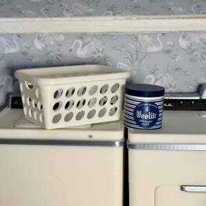 May include: A cream-colored plastic storage basket with oval cutouts and a cylindrical container of Woolite cold water wash featuring blue and white horizontal stripes and a dark blue lid, resting on top of two cream-colored appliances.