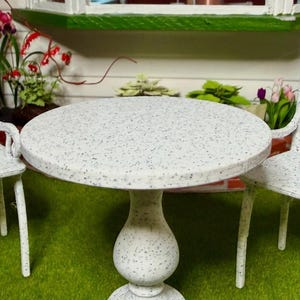 May include: A miniature round pedestal table with a white surface featuring a speckled black pattern. The table has a central column base that flares at the bottom and supports a circular tabletop. The design is consistent with dollhouse furniture.