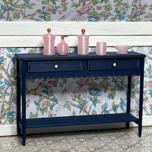 May include: A navy blue console table with two drawers and a scalloped edge, holding pink glass jars and a small bowl. The background features floral wallpaper with birds and butterflies, creating a vintage aesthetic.