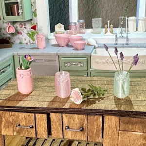 Puede incluir: Una escena de cocina en miniatura con encimera y armarios de madera. En la encimera se disponen cuencos, tarros y una maceta rosas con diseño de lazo. Un pequeño jarrón contiene flores moradas. La cocina tiene un fregadero y una ventana.