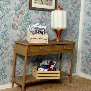 Pode incluir: Uma mesa de consola de madeira em miniatura com duas gavetas, uma borda ondulada e uma prateleira inferior. Contém um porta-revistas com revistas e uma pequena caixa de madeira com mais revistas. Uma lâmpada branca e cobre está em cima, contra um papel de parede floral.