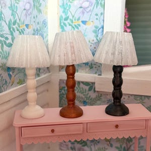 May include: Three miniature table lamps with translucent, pleated, white conical shades. The lamp bases feature a stacked, rounded design in solid white, metallic copper, and matte black finishes, standing on a pink rectangular console table.