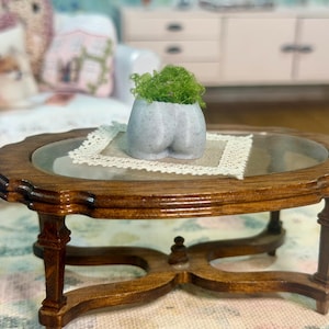 May include: A miniature, oval-shaped wooden coffee table with a glass top. A small, gray, sculpted pot with green foliage sits on a lace doily. The table has ornate legs and a dark brown finish, showcasing a vintage aesthetic.