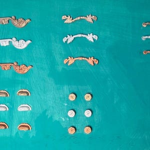 May include: An assortment of decorative hardware pieces in various metallic finishes, including gold, silver, and copper. The collection features ornate drawer pulls, handles, and decorative accents, all arranged on a teal background.