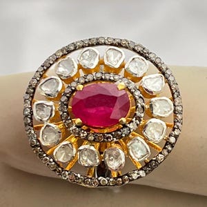 Puede incluir: Anillo dorado con un gran rubí ovalado en el centro, rodeado de piedras preciosas claras. El anillo presenta un diseño circular detallado con un borde de metal oscuro oxidado.