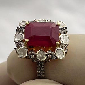 Pode incluir: Um anel de ouro e prata com uma grande pedra preciosa retangular, vermelho escuro, como peça central. A pedra preciosa é rodeada por pedras preciosas menores, claras e brancas. A banda do anel é detalhada com pequenos enfeites escuros.