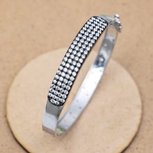 May include: A silver bracelet with a rectangular black band encrusted with small, clear gemstones. The bracelet is on a light brown surface, showcasing its polished metal and sparkling stones. The clasp is visible.
