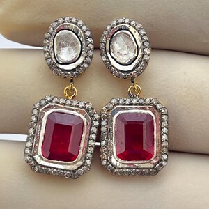 Peut inclure: Une paire de boucles d'oreilles élégantes. Chaque boucle d'oreille présente un dessus ovale avec une pierre transparente et une bordure de petits accents scintillants. En dessous, une grande pierre rouge foncé rectangulaire est sertie dans un cadre similaire.