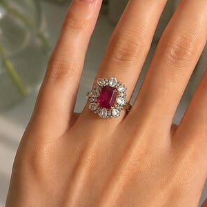May include: A ring featuring a rectangular, deep red centre stone surrounded by a halo of smaller, clear, oval-shaped stones set in a metallic band. The design showcases a prominent central gemstone framed by multiple smaller, brilliant-cut stones.