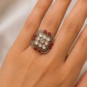 May include: A square-shaped statement ring featuring a central cluster of nine round, clear stones arranged in a grid. The centre is framed by a border of small, vibrant red stones and a delicate outer edge of tiny clear stones set in dark metal.
