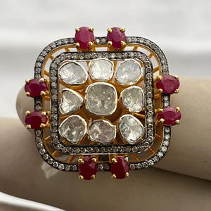 Puede incluir: Un anillo cuadrado con una banda dorada, con un grupo central de piedras claras de forma irregular. El anillo está acentuado con pequeñas piedras preciosas rojas de forma ovalada y un borde de piedras más pequeñas y brillantes.