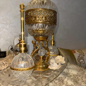 Op de afbeelding: Een sierlijke, vintage parfumfles set. De set bevat een grote, heldere glazen container met een gouden filigraan band en een kristallen top, samen met kleinere bijpassende flessen en een decoratief dienblad. De set is afgewerkt met goud metaal.