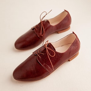 May include: A pair of burgundy leather Oxford shoes with matching laces. The shoes have a classic design with a rounded toe and a low heel. The leather appears to have a slight texture.