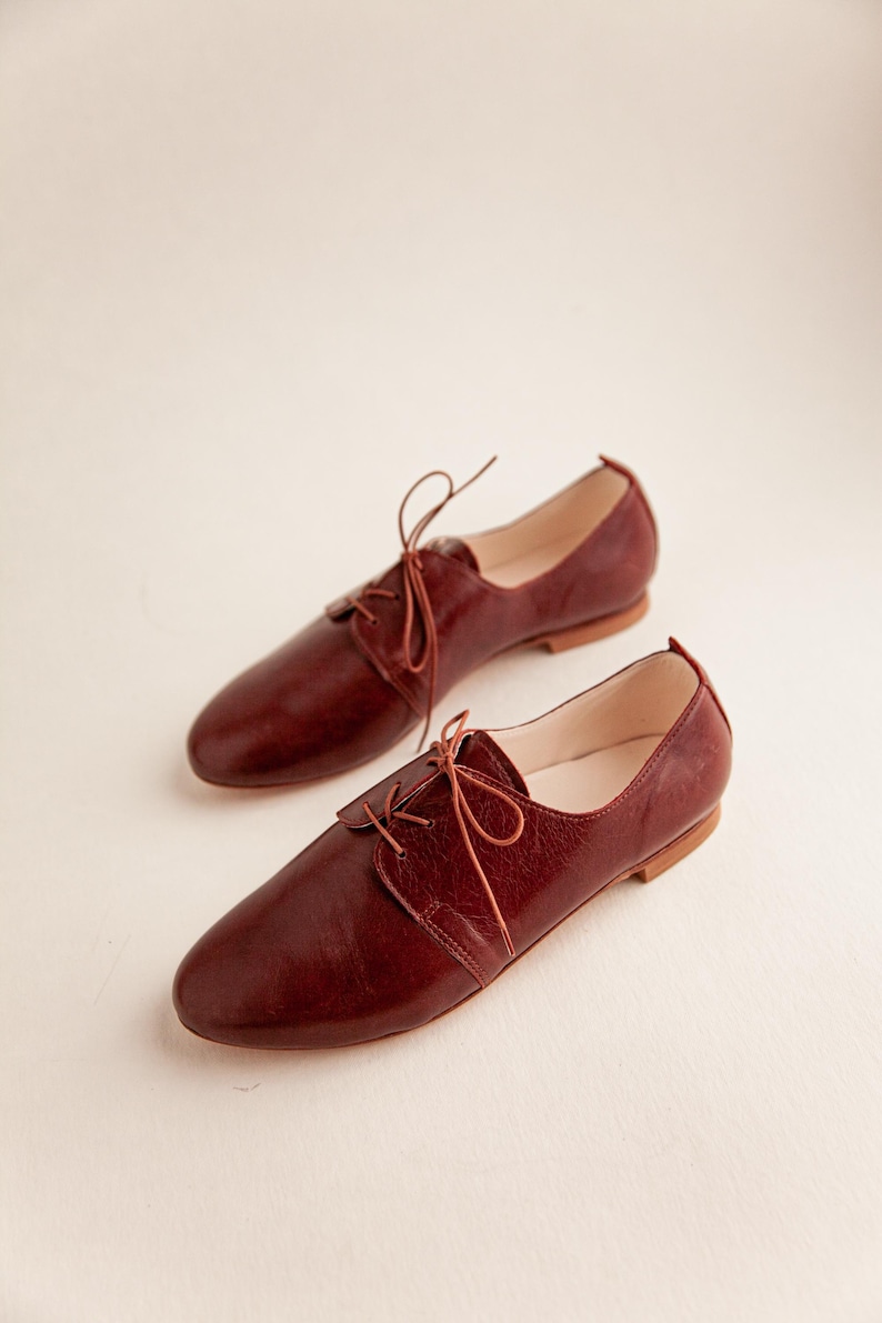May include: A pair of burgundy leather Oxford shoes with matching laces. The shoes have a classic design with a rounded toe and a low heel. The leather appears to have a slight texture.