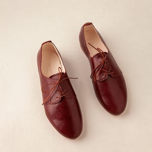 May include: A pair of burgundy leather Oxford shoes with matching laces. The shoes have a classic design with a rounded toe and a low heel. The shoes are displayed on a neutral background.