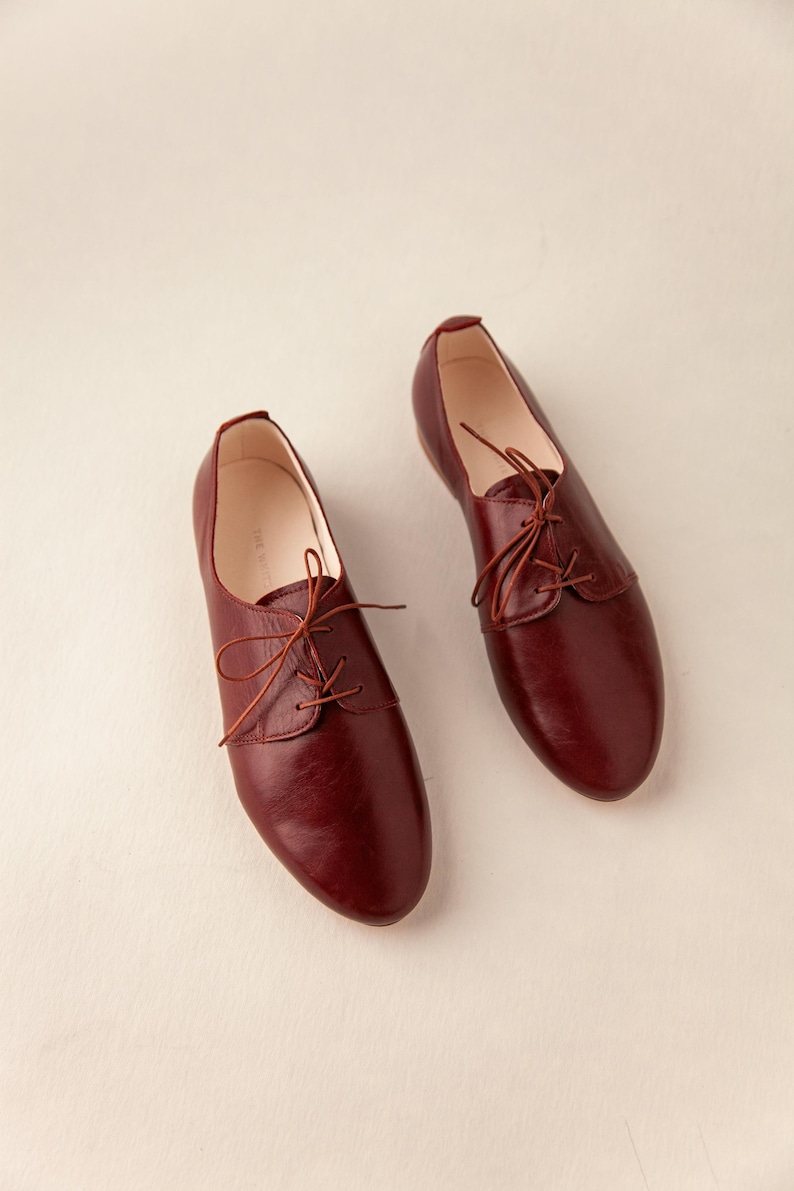 May include: A pair of burgundy leather Oxford shoes with matching laces. The shoes have a classic design with a rounded toe and a low heel. The shoes are displayed on a neutral background.