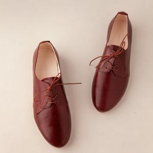 May include: A pair of burgundy leather oxford shoes with matching laces. The shoes have a classic design with a rounded toe and a low heel. The shoes are displayed on a neutral background.