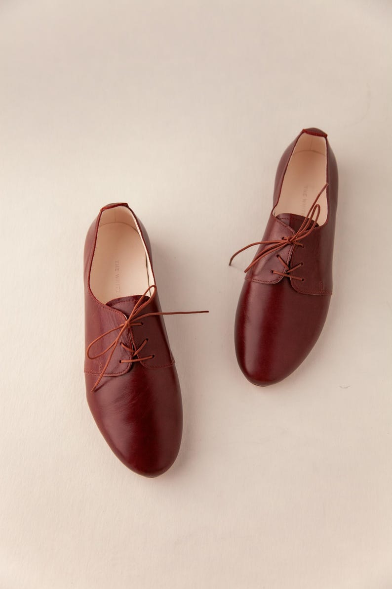 May include: A pair of burgundy leather oxford shoes with matching laces. The shoes have a classic design with a rounded toe and a low heel. The shoes are displayed on a neutral background.