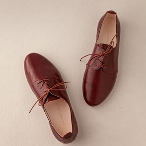 May include: A pair of burgundy leather Oxford shoes with matching laces. The shoes have a classic design with a rounded toe and a low heel. The interior of the shoe is visible, with the text "THE WHITE RIBBON" printed on the insole.